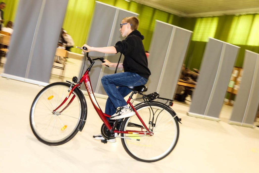 Egy fekete kapucnis pulcsit és kék farmert viselő tizenéves fiú piros biciklivel közlekedik bent. Úgy tűnik, egy kerékpáros eseményen vagy tevékenységen vesz részt egy nagy teremben, zöld falakkal és elválasztó képernyőkkel a háttérben.