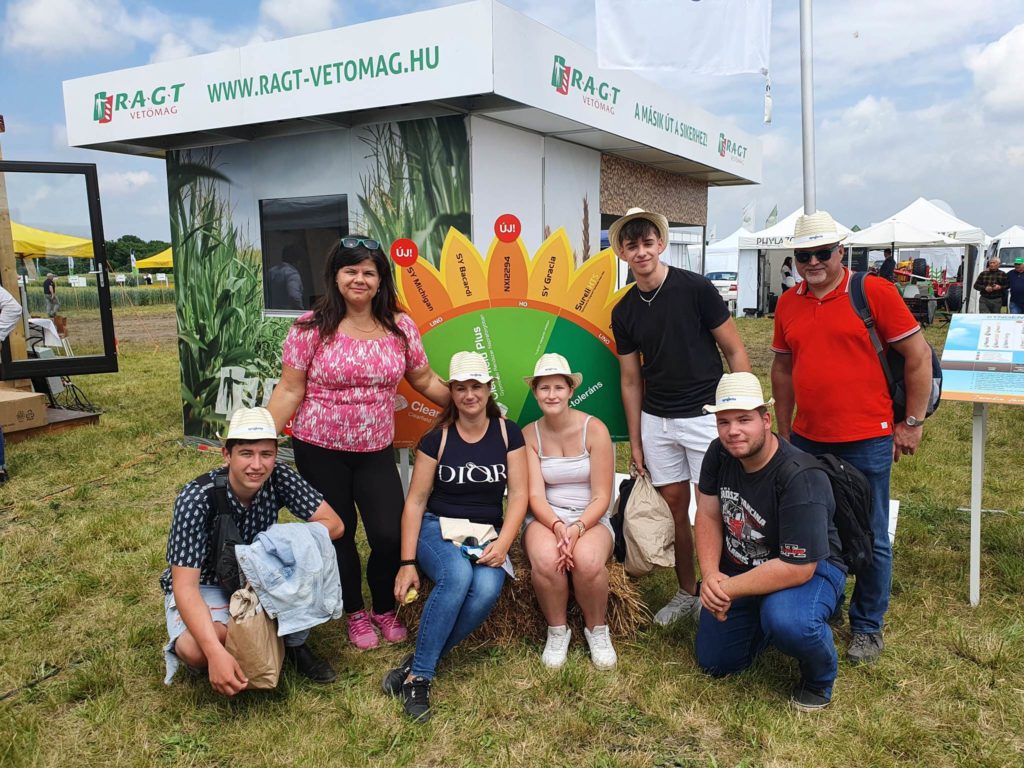 Hétfős csoport mosolyog egy szabadtéri mezőgazdasági vásári standnál, néhányan kalapot viselnek, és színes reklámháttérrel a hátuk mögött.
