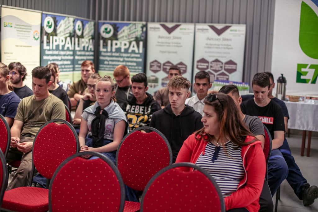 Emberek egy csoportja vörös székeken ül egy konferenciateremben, és figyelmesen hallgatja az előadást. A háttérben számos információs banner látható szöveggel és logóval. A közönség férfiakból és nőkből áll, néhányan elgondolkodtató arckifejezéssel.