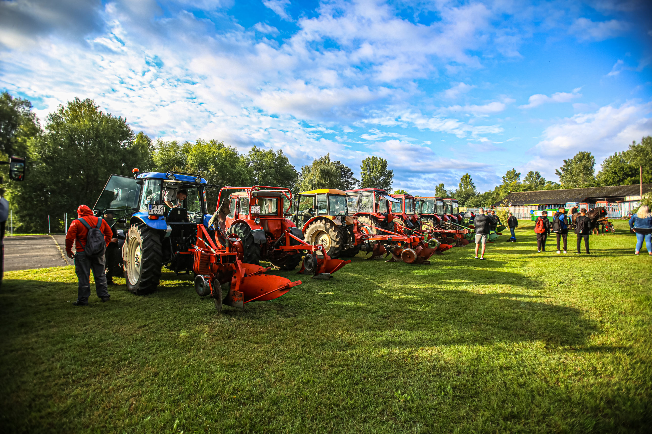 Piros és kék traktorok sora szerelvényekkel a füves mezőn, mellette egy csapat farmer gyűlt össze a kék ég alatt.