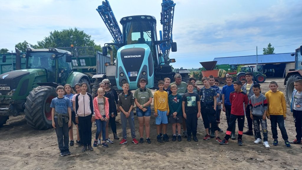 Egy csoport gyerek és néhány felnőtt nagy mezőgazdasági gépek, köztük traktorok és egy mezőgazdasági jármű előtt áll egy földön. Úgy tűnik, terepbejáráson vagy oktatási látogatáson vannak egy farmon vagy mezőgazdasági területen.