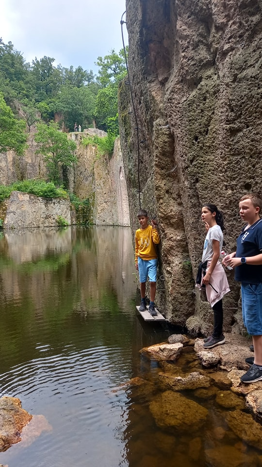 Három ember áll egy sziklás szikla mellett, nyugodt víz felett. Egy ember sárga ingben és kék rövidnadrágban hintában áll a víz felett, ugrásra készen. A többiek hétköznapi ruhában a közeli sziklákról figyelnek. Sűrű zöld fák borítják a sziklaoldalt a háttérben.