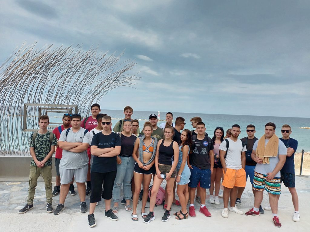 Fiatalok egy csoportja együtt pózol egy partvonal közelében. Az ég borult, a háttérben az óceán látható. A csoporttagok egy része áll, míg mások kissé a kamera felé hajolnak, a bal oldalon egy nagy fémszobor látható.