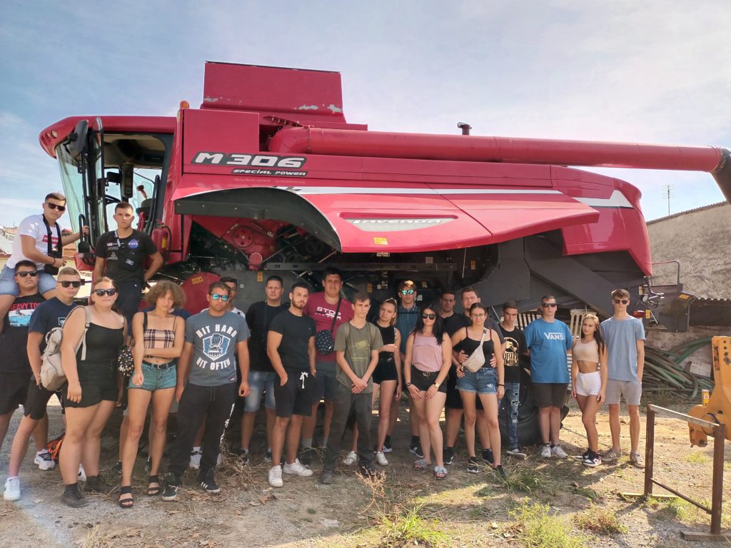 Fiatalok egy csoportja egy nagy, piros, "M306" feliratú betakarítógép előtt pózolt. A változatos, hétköznapi nyári ruhákba öltözött csapat a változóan felhős ég alatt mosolyog a kamerába. A háttérben egy vidéki, szabadtéri környezet látható mezőgazdasági berendezésekkel.