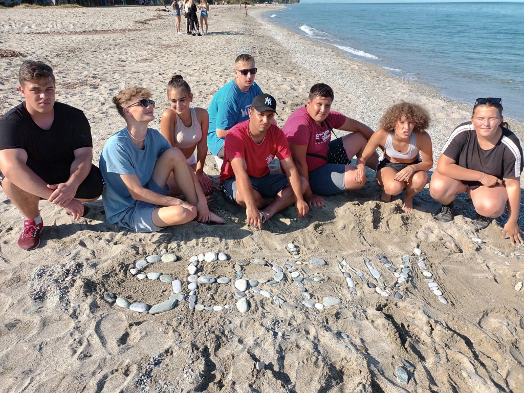 Nyolc fős csoport ül egy homokos tengerparton, közel a parthoz, nyugodt vizekkel. Fehér köveket helyeztek el a homokon, hogy kiírják maguk elé a „SERM” szót. Az ég tiszta, a háttérben mások is láthatók sétálva a tengerparton.