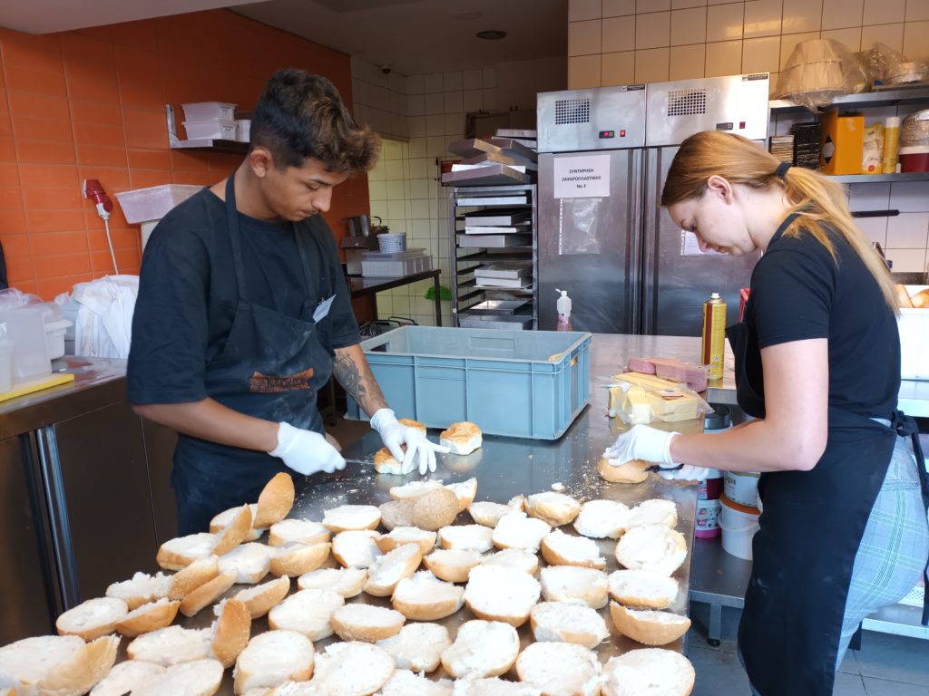 Két ember kenyeret készít egy kereskedelmi konyhában. A bal oldali személy keneget valamit a kenyérre, míg a jobb oldali a pulton lévő tárgyakat rendezgeti. Előttük a pulton számos kenyérdarab van kiterítve.