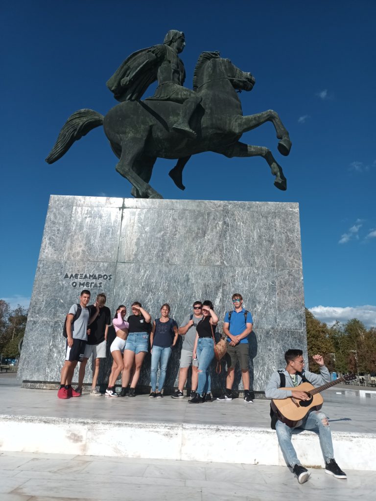 Egy csoport ember egy nagy lovas szobor előtt áll egy napsütéses napon. Vannak, akik pózolnak egy fényképen, míg egy ember a párkányon ül és gitározik. A szobor tiszta kék égen áll.
