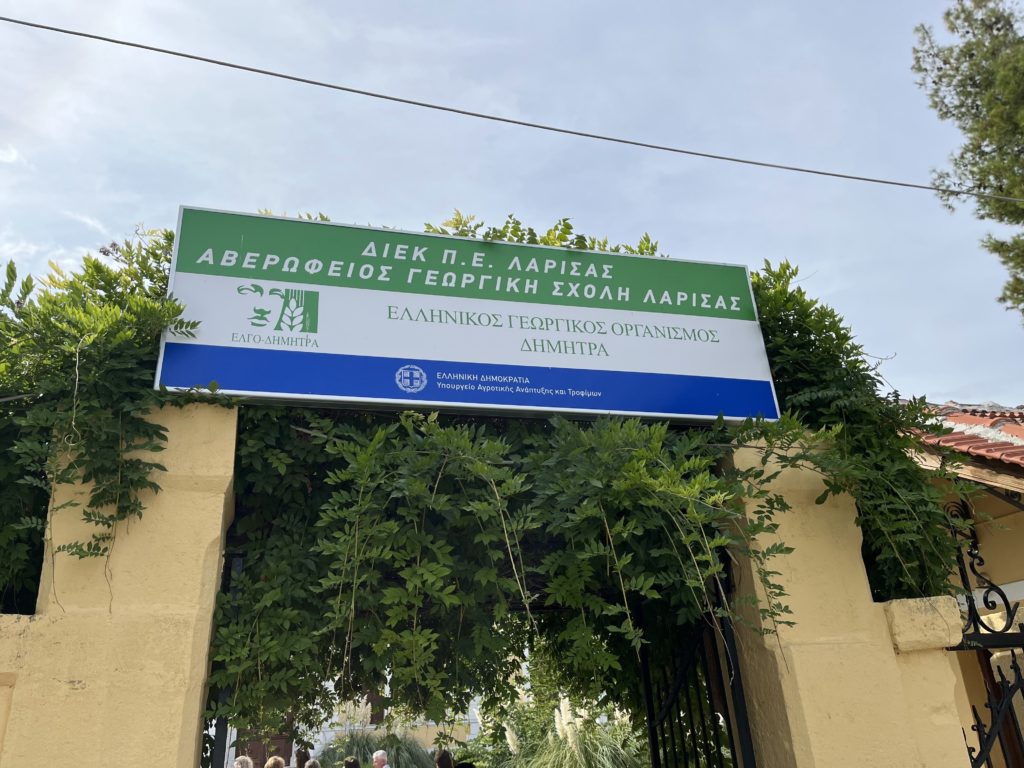 Tábla a larissai Avergios Mezőgazdasági Iskola bejáratánál, a görög DIMITRA mezőgazdasági szervezet alatt. A táblát buja zöld lombok határolják, és egy sárga kőkapu fölött található.