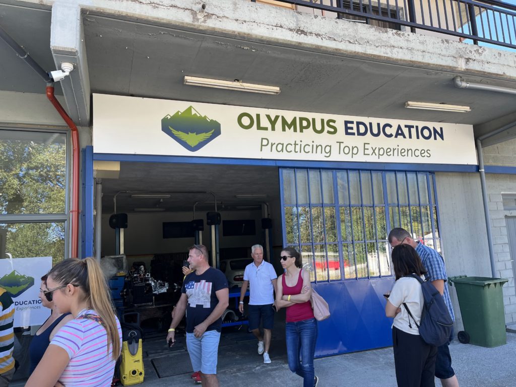 Emberek egy csoportja áll és sétál egy épület bejárata közelében, amelyen a következő felirat olvasható: „Olympus Education: Practicing Top Experiences”. Az ajtó nyitva van, felfedi benne a berendezés. Az épülethez tágas felső erkély tartozik.