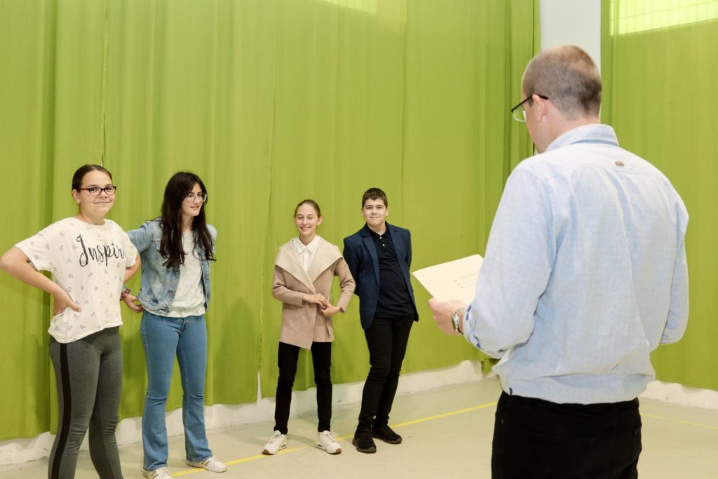 Egy tanár négy diákkal áll szemben, akik sorakoznak a zöld függönyök hátterében. Úgy tűnik, hogy a tanulók vitában vagy tevékenységben vesznek részt, az egyik diák csípőre tett kézzel pózol, míg a többiek figyelmesen hallgatják.