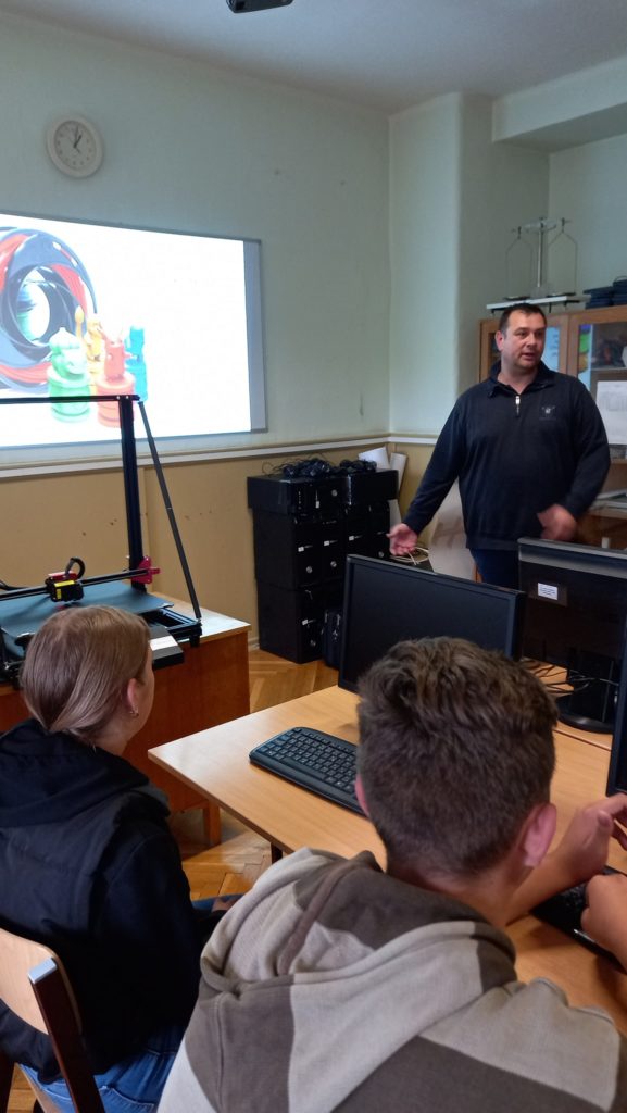 Egy osztálytermi jelenet egy férfi tanárral, aki a számítógép és a kivetítő képernyője mellett áll, színes grafikákat megjelenítve. három diák néz szembe vele, figyelmesen hallgatva.