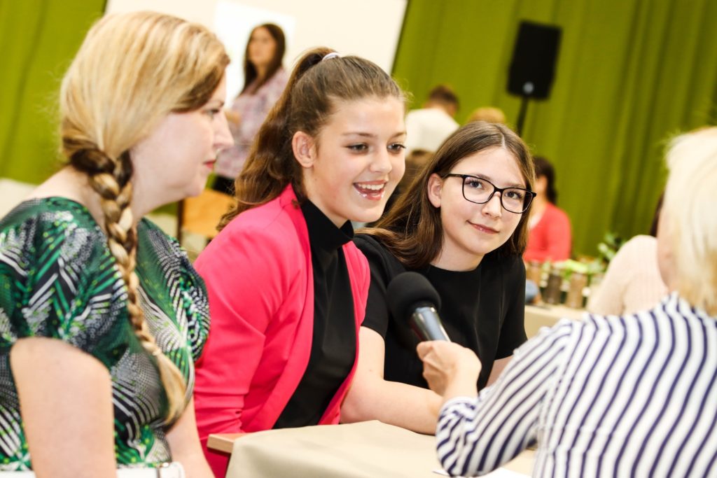 Három fiatal nő ül egy asztalnál, és egy mikrofont tartó személy interjút készít velük. A középen álló, rózsaszín kabátot viselő nő beszél. A mellette lévő nőnek hosszú copfoja van, a másik nő szemüveges. A háttérben zöld függöny látható.