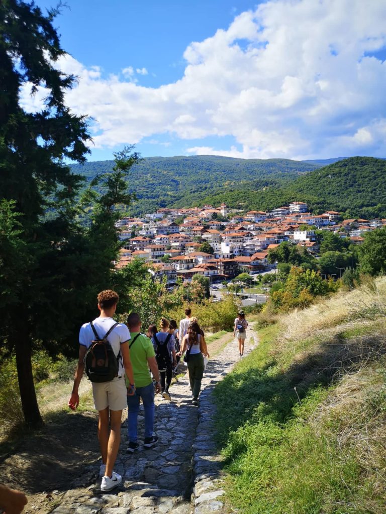 Emberek egy csoportja egy kőösvényen sétál egy festői falu felé, amely buja zöld dombok között fészkel, a fényes kék égbolyhos felhők alatt. A falut vörös cseréptetők borítják, és erdős hegyek veszik körül.