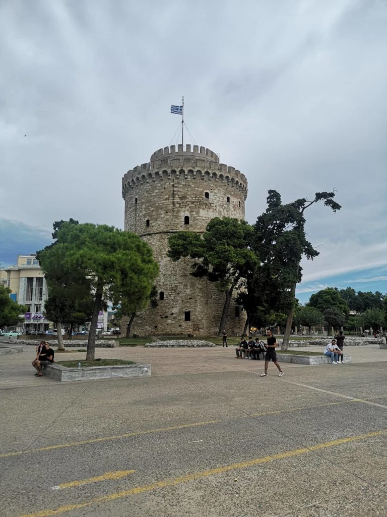 A képen Thesszaloniki Fehér Toronya látható, egy kerek kőtorony, tetején görög zászlóval. Fák, padok és sétáló vagy ülő emberek veszik körül. Az ég borús, és a torony jól látható egy nyílt nyilvános térben.