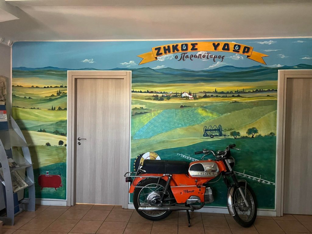Egy régi, narancssárga motorkerékpár fekete üléssel egy falfestmény mellett parkolt beltérben. A falfestmény hullámzó vidéki tájat ábrázol mezőkkel, dombokkal és egy távoli faluval. Fent egy szalaghirdetésen görög nyelvű szöveg található. A háttérben két zárt ajtó látható.