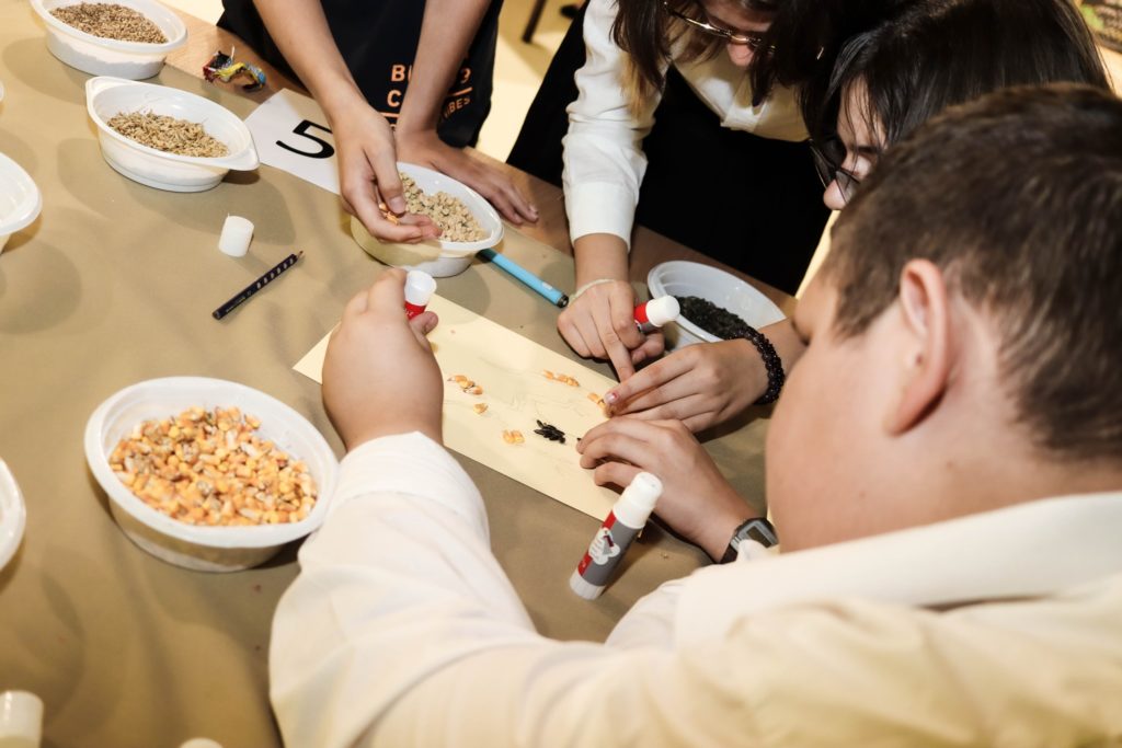Diákok egy csoportja összegyűlik egy asztal körül, akik egy kézműves projekten dolgoznak. Ragasztópálcákat használnak arra, hogy tálakból apró tárgyakat, például magvakat és szemeket rögzítsenek a papírra. Az asztalt különféle tálakba rendezett kellékek borítják.
