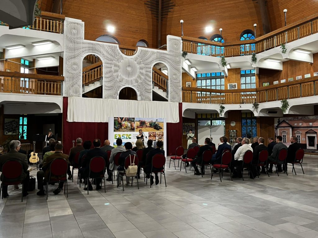 Beltéri rendezvény egy fából készült belső térben, ahol piros székek fehér függönyös színpad felé néznek. emberek ülnek, és két hangszórót figyelnek egy „keleti” feliratú transzparens közelében.