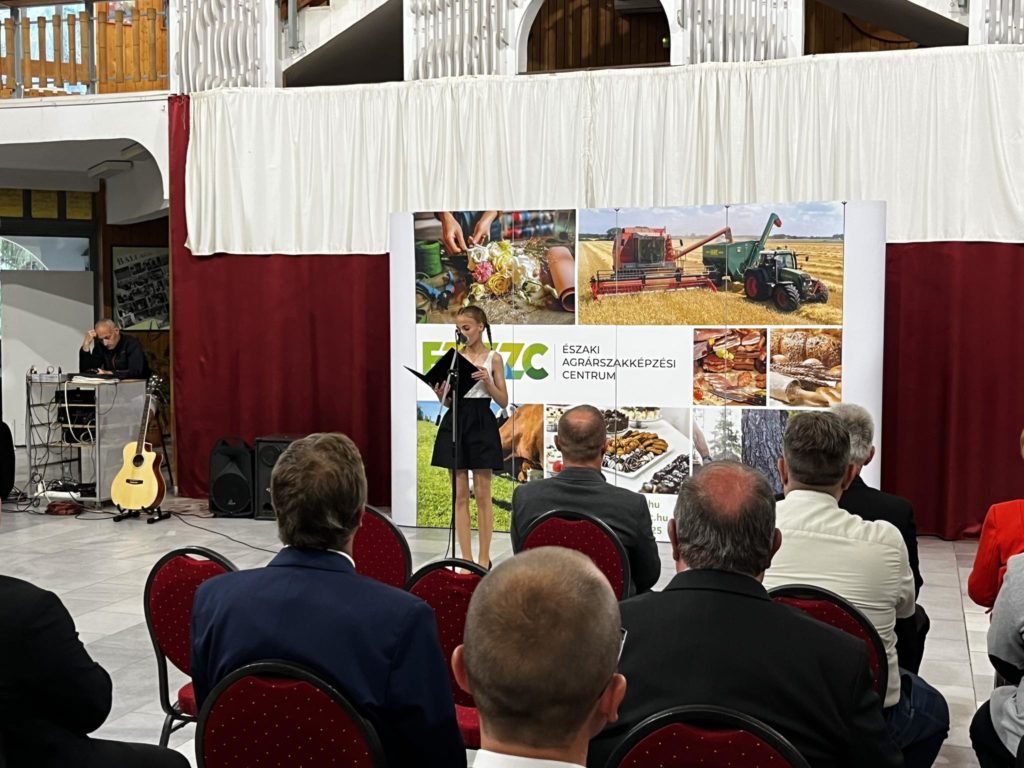 Egy nő beszél a pódiumon a közönség előtt, és egy nagy mezőgazdasági témájú poszter egy konferenciateremben, piros székekkel és egy zenésszel az oldalán.