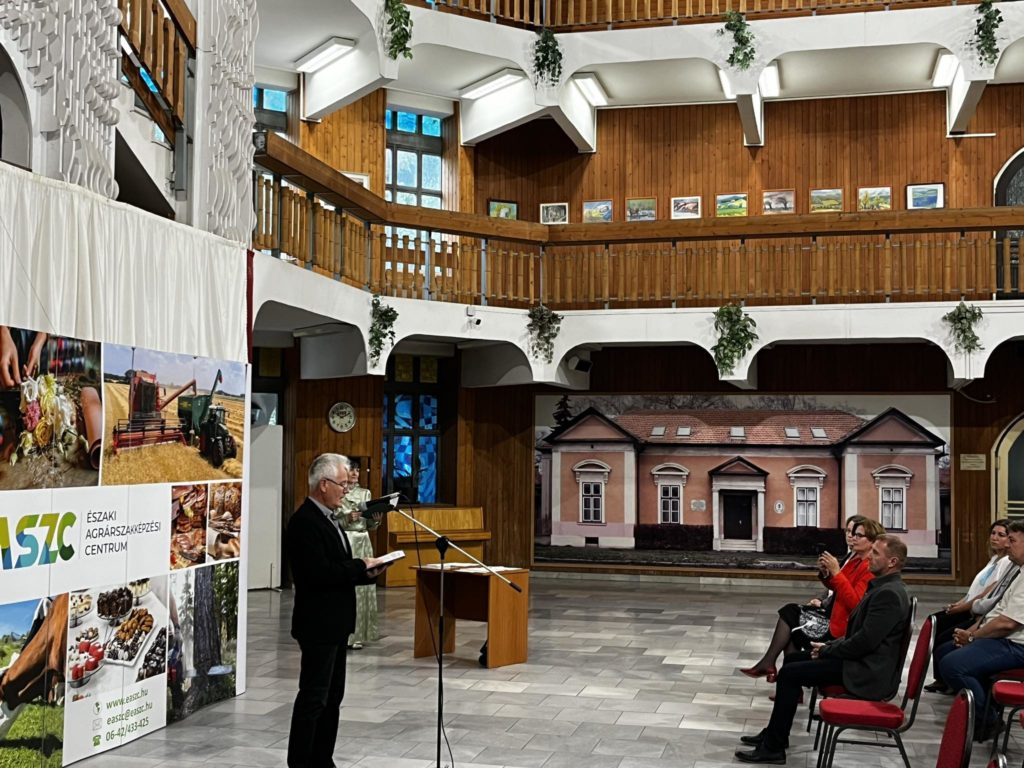 Egy férfi beszél egy pódiumon egy erkélyszintes, fából készült építészettel és ülő résztvevőkkel rendelkező teremben. mezőgazdasági képekkel ellátott transzparensek lógnak a háttérben.