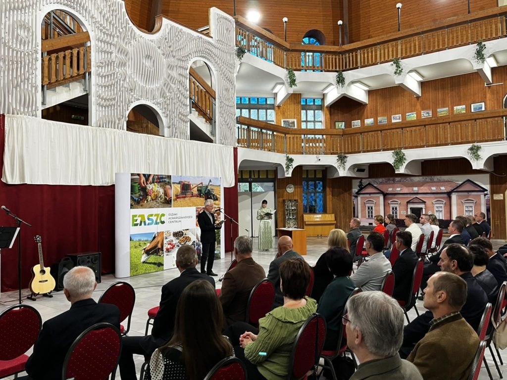 Egy előadó szól a közönséghez egy nagy faerkélyekkel és kulturális dekorációval rendelkező teremben, egy rendezvényen mezőgazdasági képeket bemutató transzparensekkel.