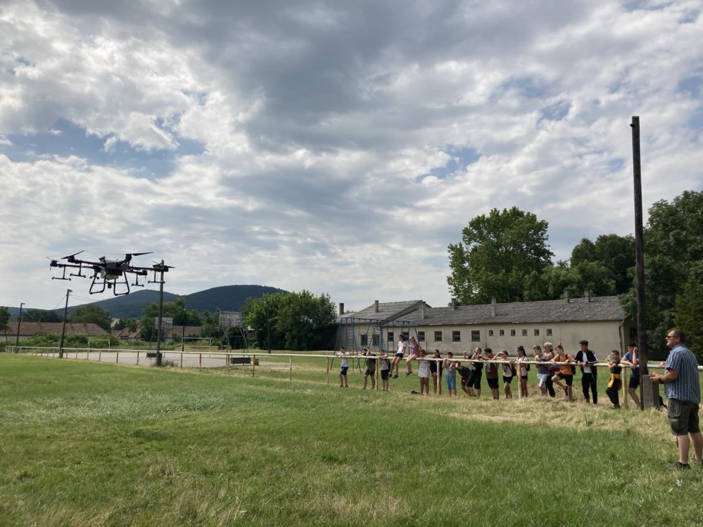 Emberek nagy csoportja egy drónt néz a szabadban. A jelenet egy füves területen játszódik, felhős égbolttal és épületekkel a háttérben. Úgy tűnik, hogy a csoport, némelyik kamerát tart, egy drónbemutatót vagy eseményt figyel meg vagy vesz részt rajta.