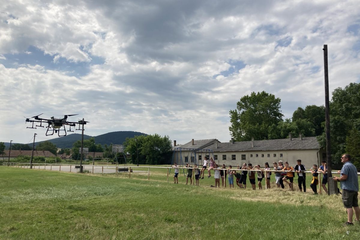 Egy csoport ember összegyűlik a szabadban egy füves mezőn, hogy egy nagy drónt nézzenek repülés közben. Felhős az ég, a háttérben épületek és dombok láthatók. Egy személy az előtérben egy irányítót tart, valószínűleg a drónt irányítja.