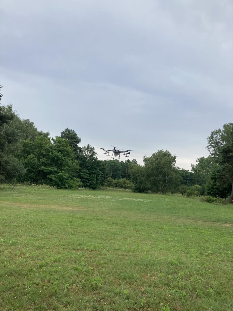 Egy nagy drón több rotorral repül egy fákkal körülvett füves mező felett a borús égbolt alatt.
