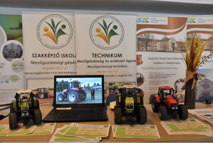 A mezőgazdasági technológiát bemutató standon három traktormodell, egy traktorvideót bemutató laptop és számos brosúra mutat be. A háttérben transzparensek egy mezőgazdasági és erdészeti szakképző iskolát és képzési programokat hirdetnek.