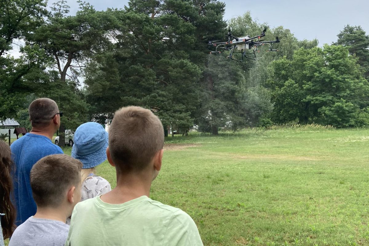 Gyerekek és felnőttek egy csoportja egy füves mezőn áll, és egy drónt néz. A drón a levegőben lebeg fákkal és felhős égbolttal a háttérben. Úgy tűnik, hogy a jelenet szabadtéri tevékenység vagy bemutató közben játszódik.