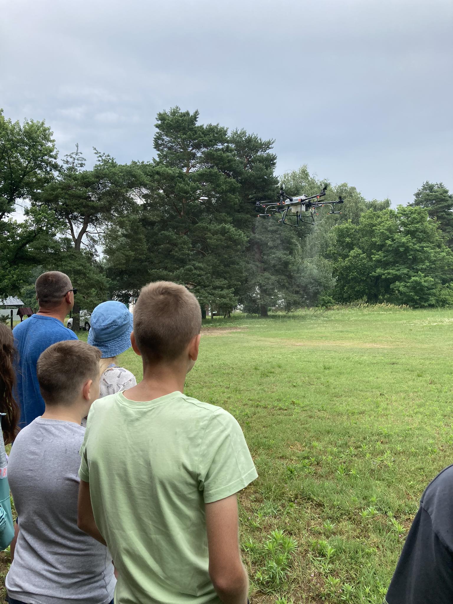 Gyerekek és felnőttek egy csoportja egy füves mezőn áll, és egy drónt néz. A drón a levegőben lebeg fákkal és felhős égbolttal a háttérben. Úgy tűnik, hogy a jelenet szabadtéri tevékenység vagy bemutató közben játszódik.