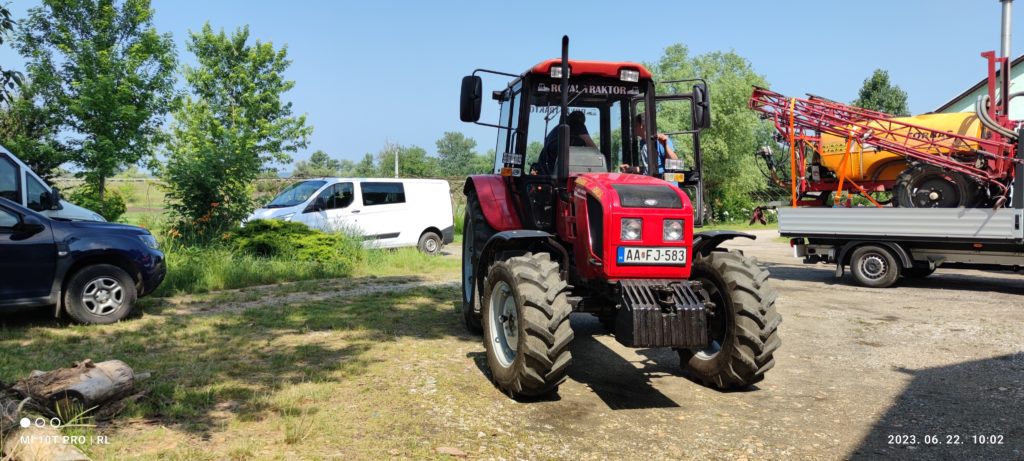 Egy élénkpiros, "AAFJ 538" rendszámú traktor parkolt egy fűvel és fákkal körülvett földúton. Két kisteherautó, egy fekete és egy fehér, látható a háttérben, valamint néhány mezőgazdasági berendezés egy pótkocsin. A fénykép 2023. június 22-i keltezésű.
