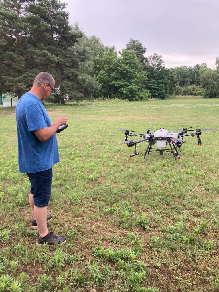 Egy férfi áll egy füves területen, és egy nagy drónt üzemeltet egy távirányítóval. A több rotorral és érzékelővel felszerelt drón a földön fekszik előtte. A háttérben fák és felhős égbolt látható.