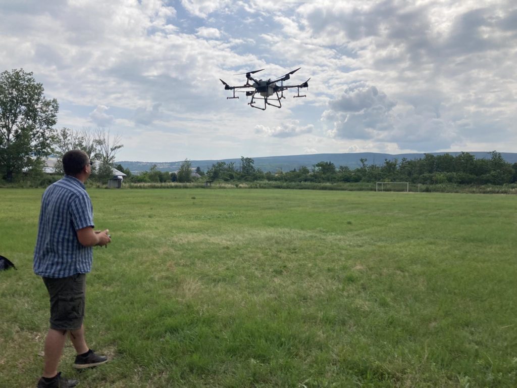 Egy férfi egy nagy drónt üzemeltet egy nyílt füves területen, néhány fával a távolban. Az ég részben felhős, a tájat enyhe dombok jellemzik. A férfi csíkos inget és rövidnadrágot visel, és a drón irányítására összpontosít.