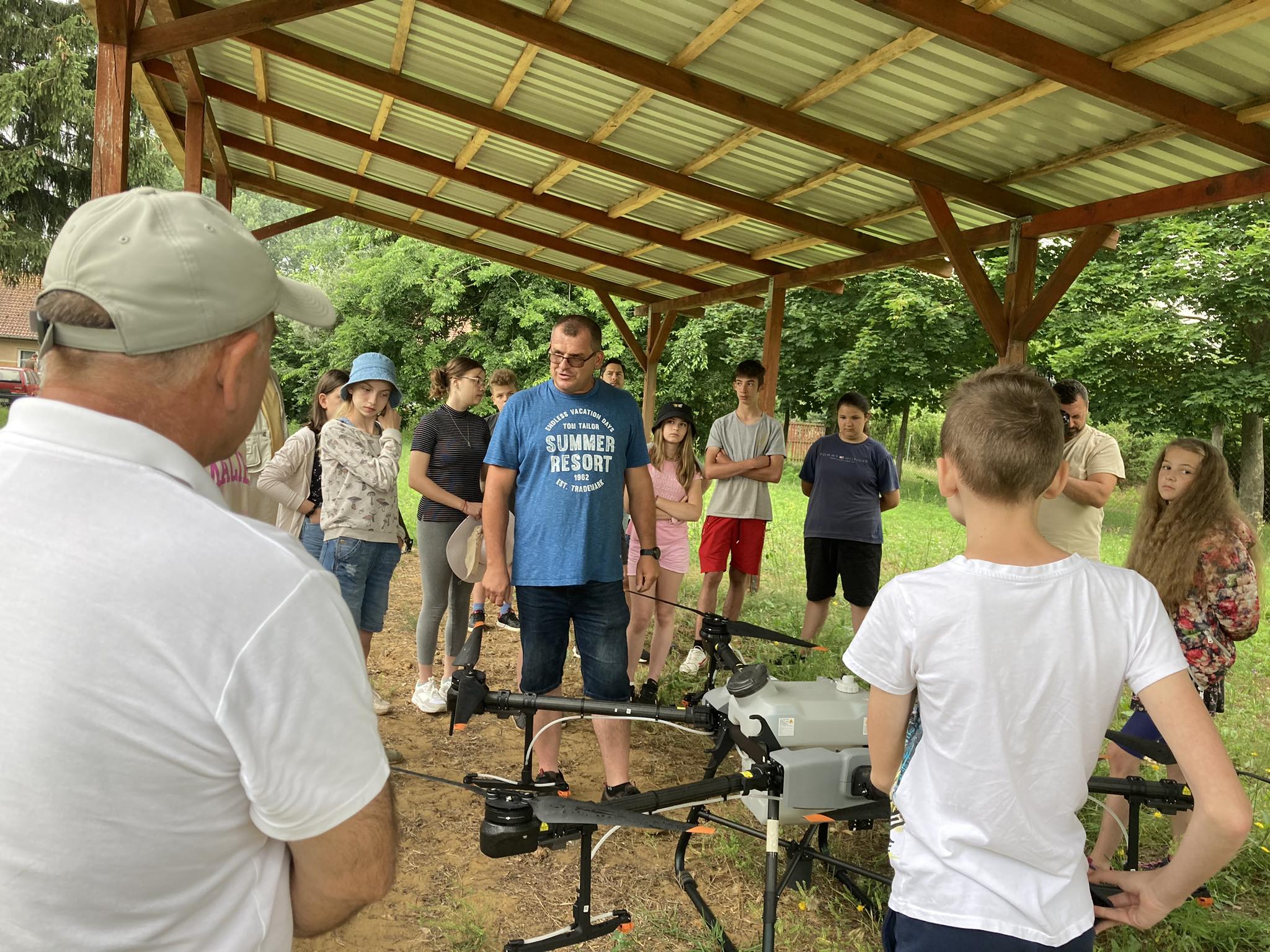 Egy csapat ember egy fából készült lombkorona alatt áll egy kertben, egy kék pólós férfi köré gyűlve, aki a jelek szerint a földön lévő drónról magyaráz valamit. A csoport felnőttekből és gyerekekből áll, mindannyian figyelmesen hallgatnak.