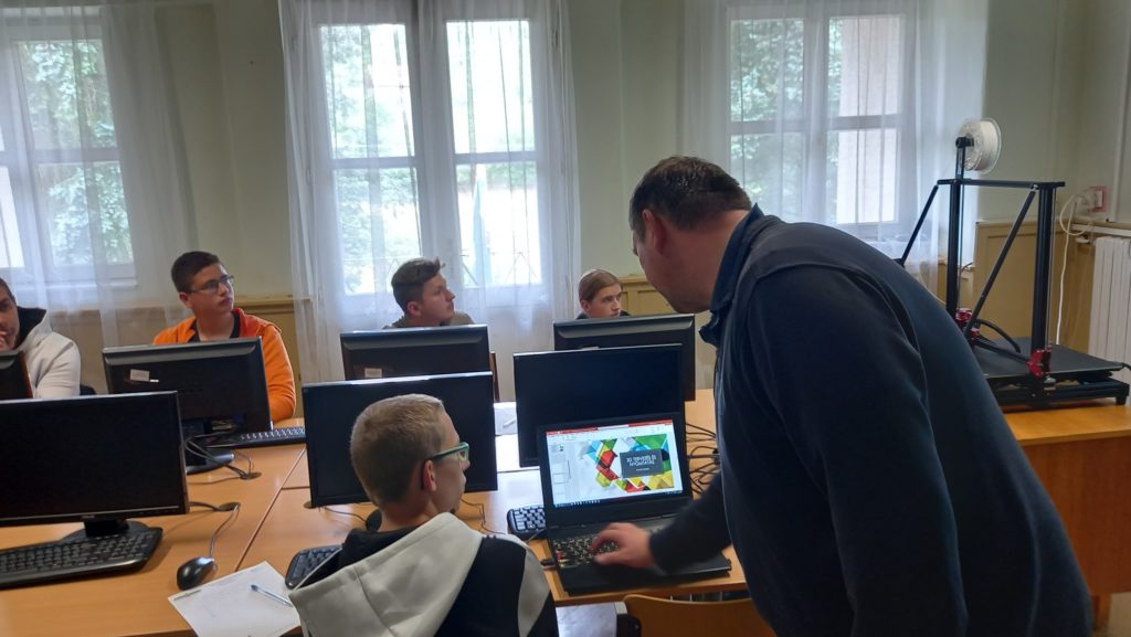 Egy tanár számítógéppel segíti a diákot, míg négy másik diák figyelmesen figyel az osztályteremben. A beállítás több számítógépet tartalmaz egy nagy fából készült asztalon és a háttérben lévő ablakokat, amelyek természetes fényt engednek be.