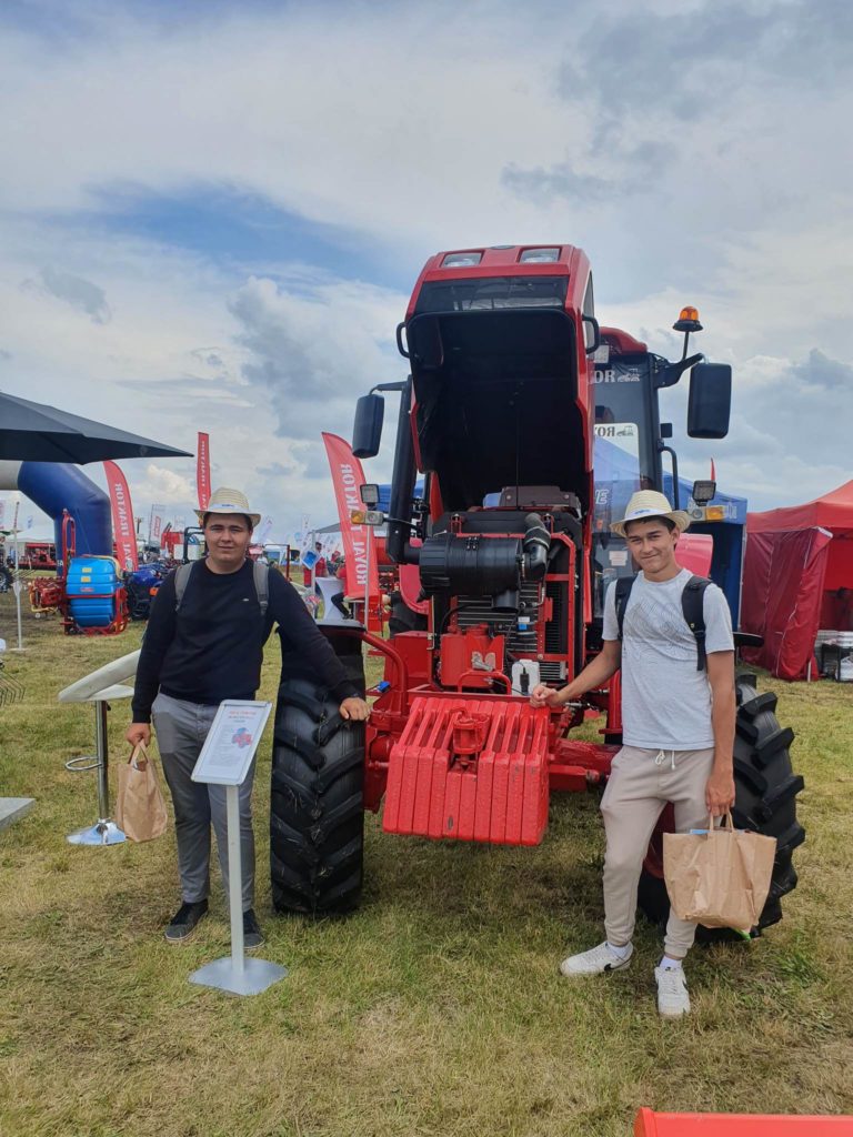 Két férfi állt egy nagy piros traktor előtt egy szabadtéri kiállításon. mindegyik férfi egy táskát tart a kezében, és mosolyog a kamerába, a háttérben különféle egyéb gépek és sátrak láthatók.