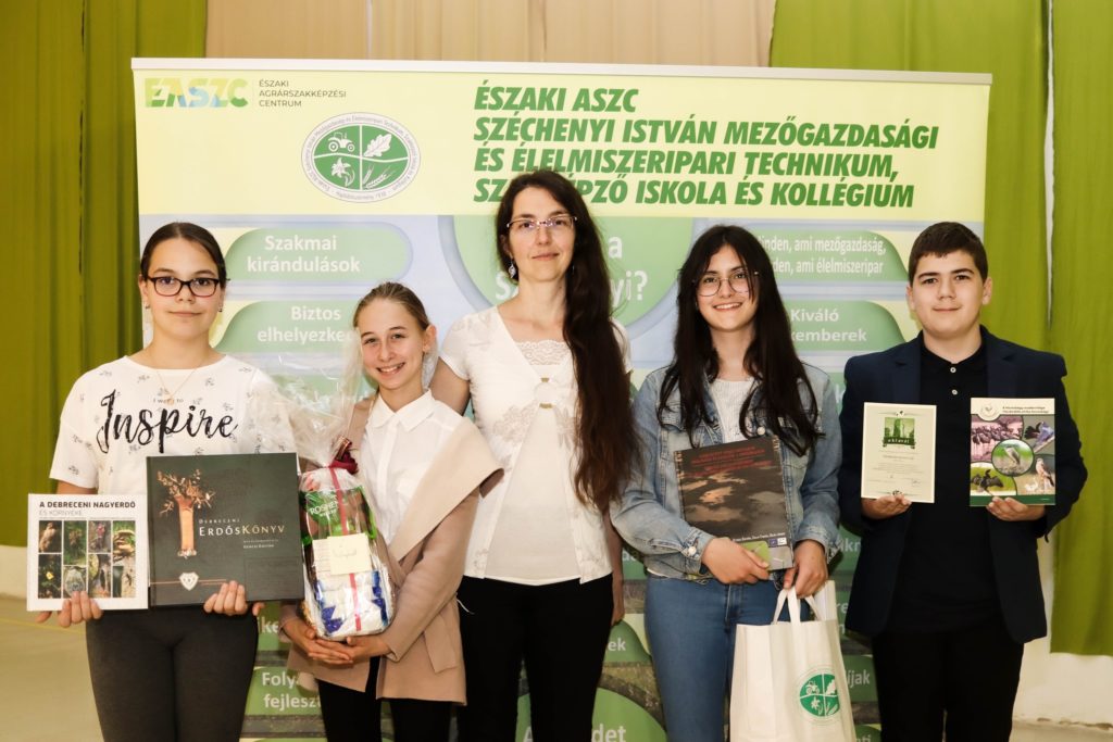 Öt ember áll egy mezőgazdasági és élelmiszertechnológiai oktatási központ reklámszalagja előtt. A négy fiatal egy könyvet és bizonyítványt tart a kezében, amelyek bemutatják eredményeiket. A mögötte lévő transzparens magyar nyelvű információkkal szolgál az oktatási központról.