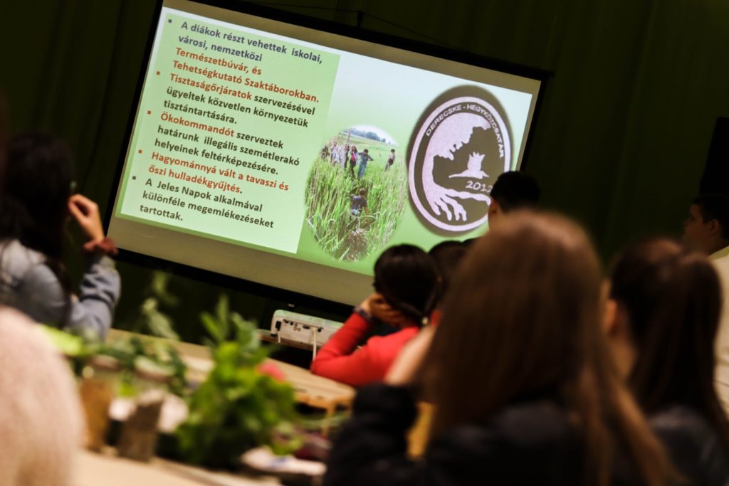 Emberek egy csoportja ül egy szobában, szemben a szöveget és képeket megjelenítő prezentációs képernyővel. A dián a környezetvédelmi tevékenységeket tárgyaló magyar nyelvű pontok listája, kör alakú logó és egy szabadtéri környezetben lévő emberek fényképe látható.