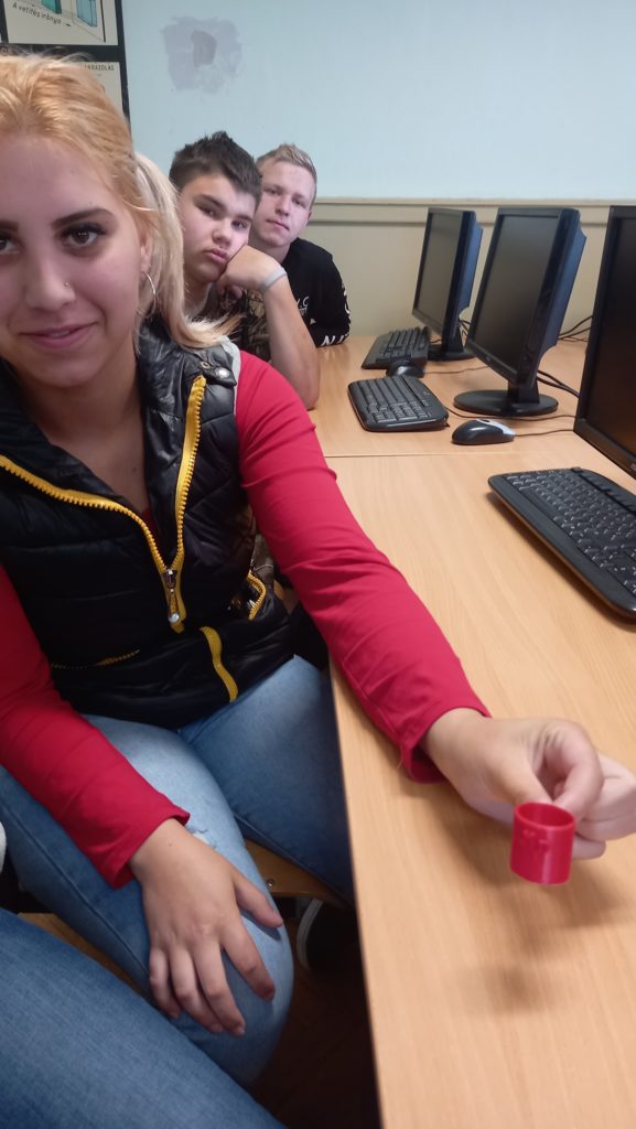Egy szőke hajú, piros inget és fekete mellényt viselő fiatal nő kis piros sapkát tart egy faasztalnál az osztályteremben. Két másik ember, egy fiatal férfi és egy fiatal nő ül mögötte, számítógépekkel az asztalokon.