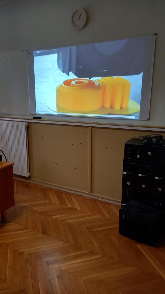 Egy osztályterem egy nagy digitális képernyővel, amelyen egy szeletelt narancs és egy csésze tea képe látható, egy egyszerű fehér fal mellett, fapadlóval és egy fekete tárolószekrénnyel a jobb oldalon.