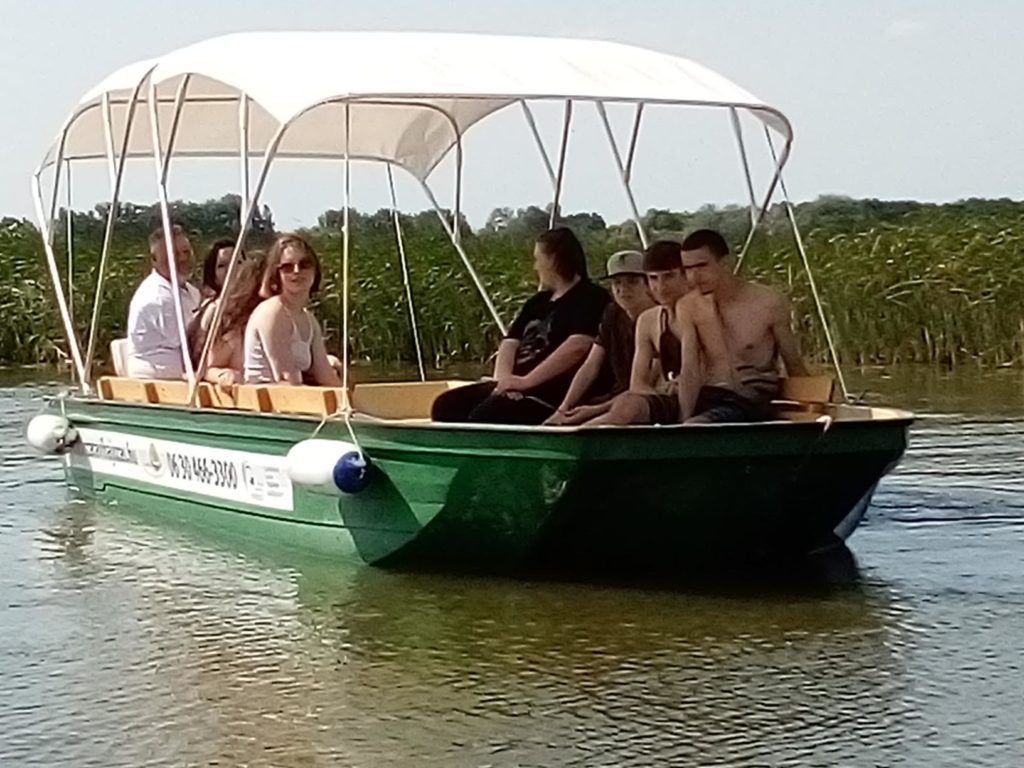 Emberek csoportja ül egy motorcsónakon fehér lombkoronával, lebeg egy nyugodt vízen. A csónak zöld színű, és egy elérhetőségi szám látható az oldalán. A háttérben nád és tiszta égbolt látható.