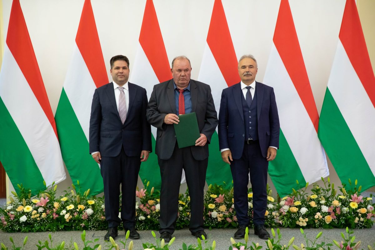 Három öltönyös férfi áll egymás mellett egy zászlóra emlékeztető, váltakozó piros, fehér és zöld csíkokkal díszített háttér előtt. Virágkompozíciókat helyeznek a lábuk elé. A középső férfi egy zöld mappát tart a kezében.