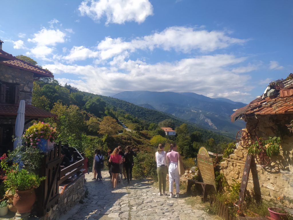 Festői hegyi falu kőösvényekkel és rusztikus épületekkel. Az előtérben egy csoport ember sétál a zöld dombokra és hegyekre néző kilátó felé, részben felhős égbolt alatt. Virágok és növények díszítik az ösvény széleit.