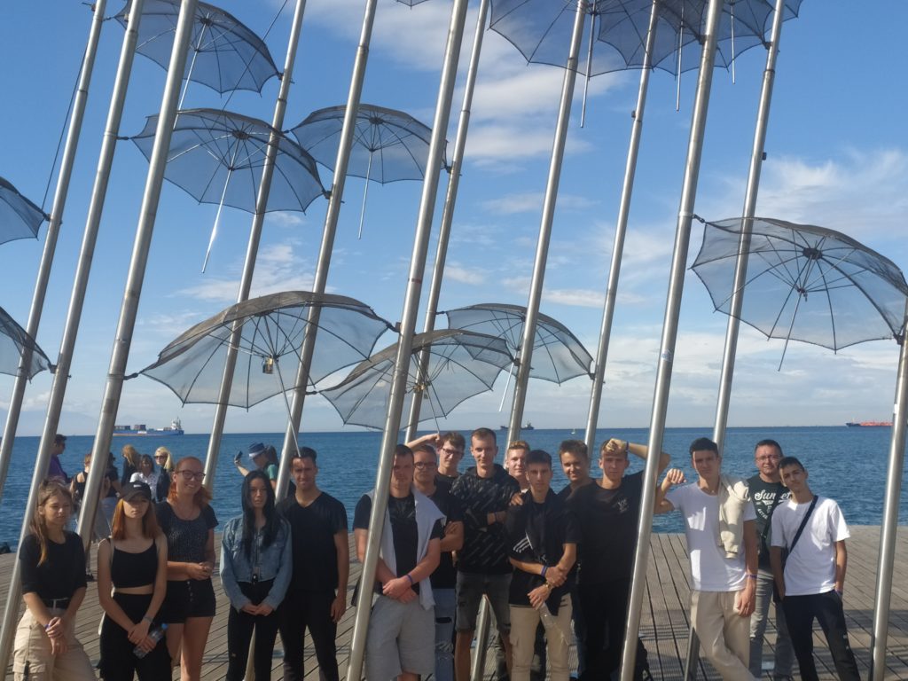 Emberek egy csoportja áll egy szabadtéri művészeti installáció előtt, amelyen több fémesernyőszobor látható a tiszta kék ég alatt. Az óceán látható a háttérben, és néhány hajó látható a horizonton. Úgy tűnik, hogy a csoport egy fényképhez pózol.