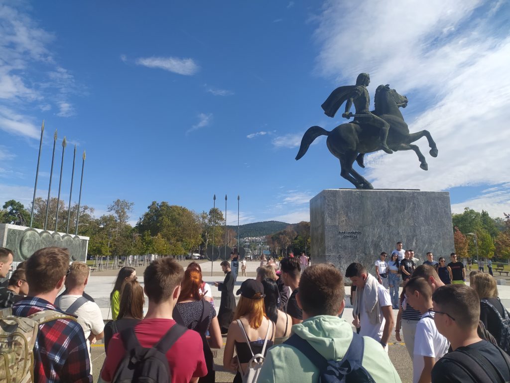 Egy csoport ember gyűlik össze egy nagy lovas szobor előtt, ragyogó kék ég alatt. A szobor betontalapzaton áll, a háttérben fák és zászlórudak láthatók. Az emberek turistáknak vagy egy szabadtéri rendezvény résztvevőinek tűnnek.
