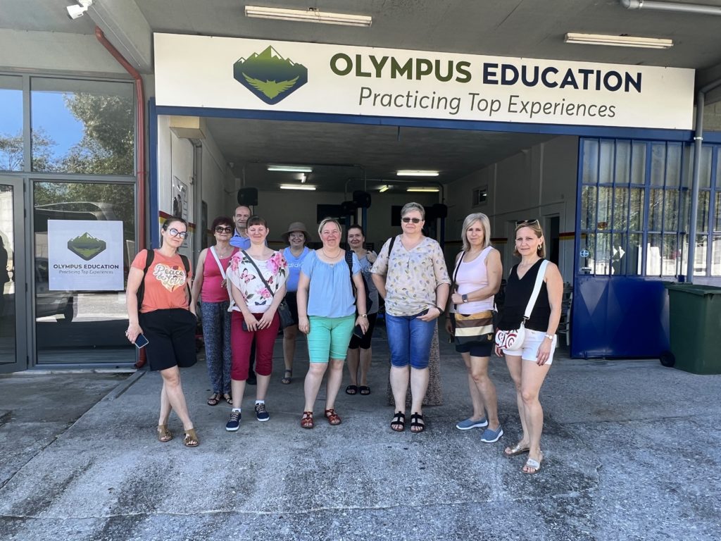 Egy tizenegy emberből álló csoport áll egy épület előtt, amelyen az „Olympus Education: Practicing Top Experiences” felirat olvasható. Könnyen öltözködnek, és néhányan mosolyognak. Az épület nagy ablakokkal és üvegajtóval rendelkezik.