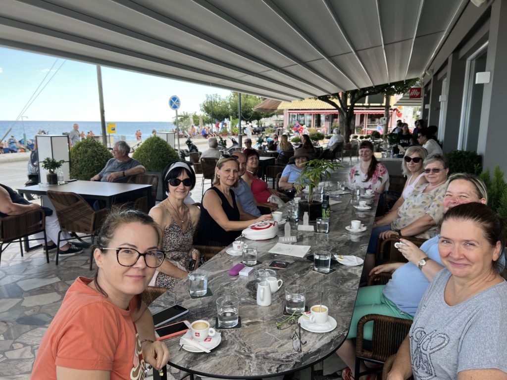 Egy csapat ember ül egy hosszú asztal körül egy szabadtéri kávézóban a strand közelében, és mosolyog a kamerába. Az asztalon számos kávéscsésze és egy kis sütemény található. A háttérben más látogatók és az óceánra néző kilátás látható.