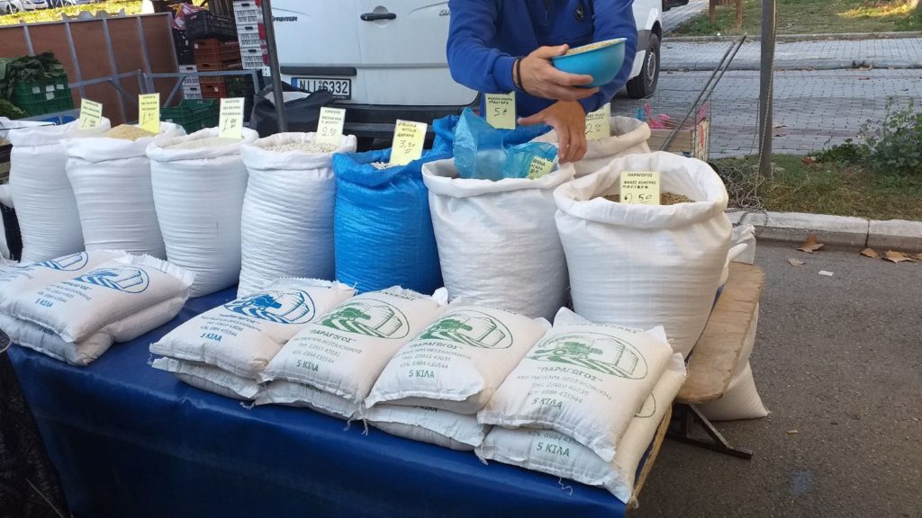 Egy piaci standon különféle rizsfajták és gabonafélék láthatók nagy fehér és kék zsákokban, mindegyiken feltüntetve az árat. Néhány kisebb táska zöld és kék jelzéssel szépen elrendezve az alábbi táblázatban. Egy személy kikanala valamit az egyik zsákból.