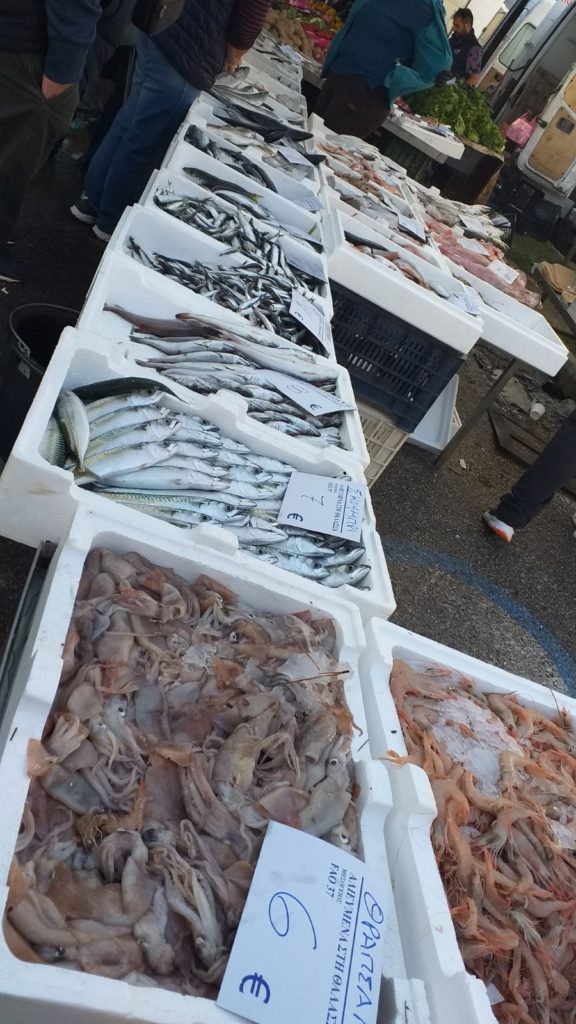 A tenger gyümölcseit kínáló piac standján különféle friss halak és tenger gyümölcsei, köztük tintahal, garnélarák és különféle halak találhatók, fehér habtartályokba rendezve. Egyes konténereken eurós árcédulákat helyeztek el. Az emberek a háttérben vásárolnak.