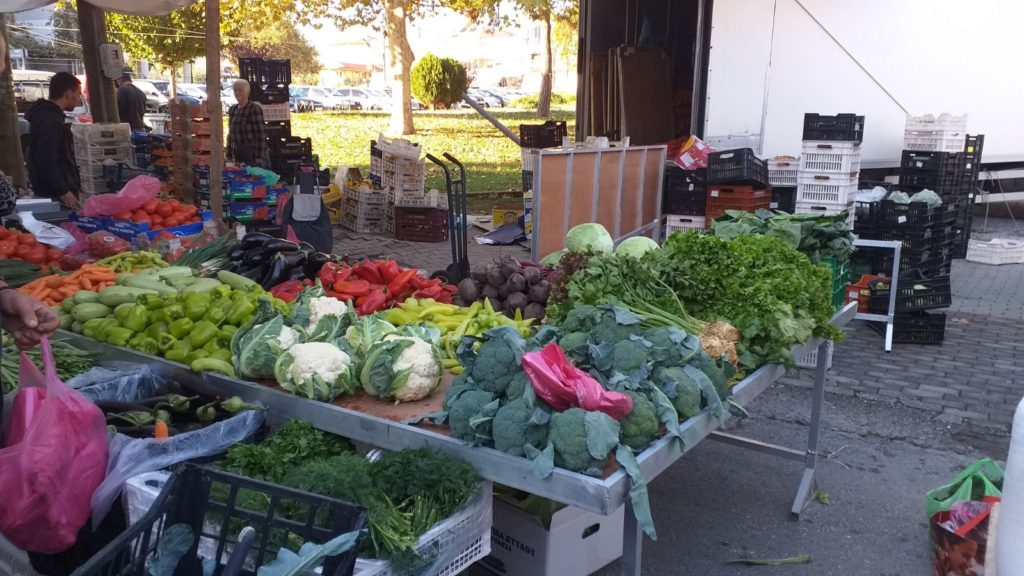 A nyüzsgő szabadtéri piaci standon számos friss zöldség található, köztük karfiol, leveles zöldek, sárgarépa, paprika, cékla és káposzta. A háttérben bevásárló emberek láthatók, a bódét pedig dobozok és egyéb piaci kellékek veszik körül.